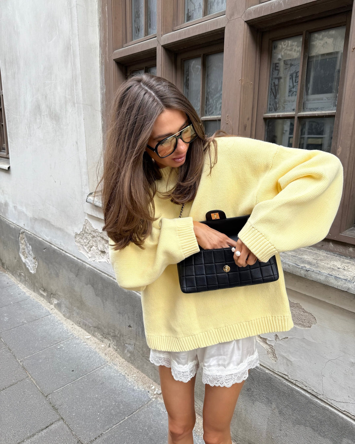 Woman wearing a yellow Letter-print Loose-fit Sweater, holding a black clutch, posing stylishly outdoors.
